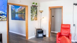 A room with a wooden floor, a red chair, a painting of a lake, a lamp, and a small table with a laptop.
