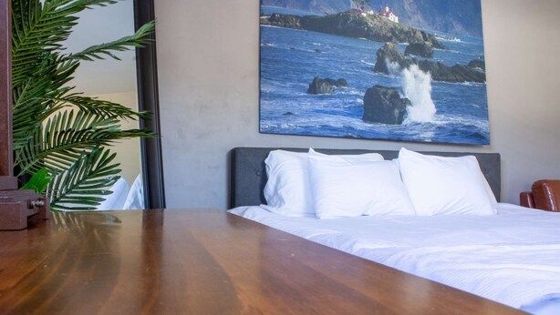 A bedroom with a wooden bed, white bedding, a large painting of a coastal scene, and a potted plant.