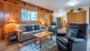 Ein gemütliches Wohnzimmer mit einer Holzverkleidung, einem Sofa, einem Couchtisch, einem Ledersessel und einem Fenster mit Blick auf Bäume.