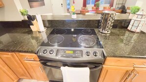 Cocina con estufa negra, horno y encimera de granito. Cuenta con alacenas de madera y una ventana con cortinas.