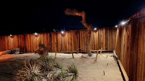 Un área exterior cercada con suelo arenoso, valla de madera y algunas plantas propias del desierto.