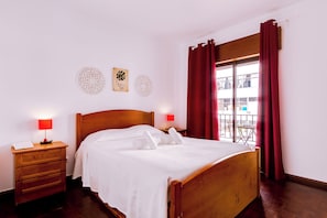 A neatly made bed with white linens, a wooden headboard, and two bedside lamps with red shades. - Anya (FARO)