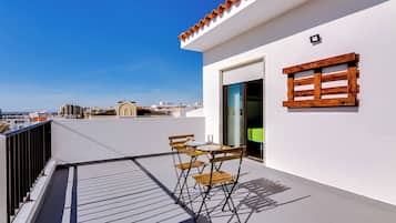 Un balcón con mesa y sillas, una ventana con contraventanas de madera y vista a la ciudad.