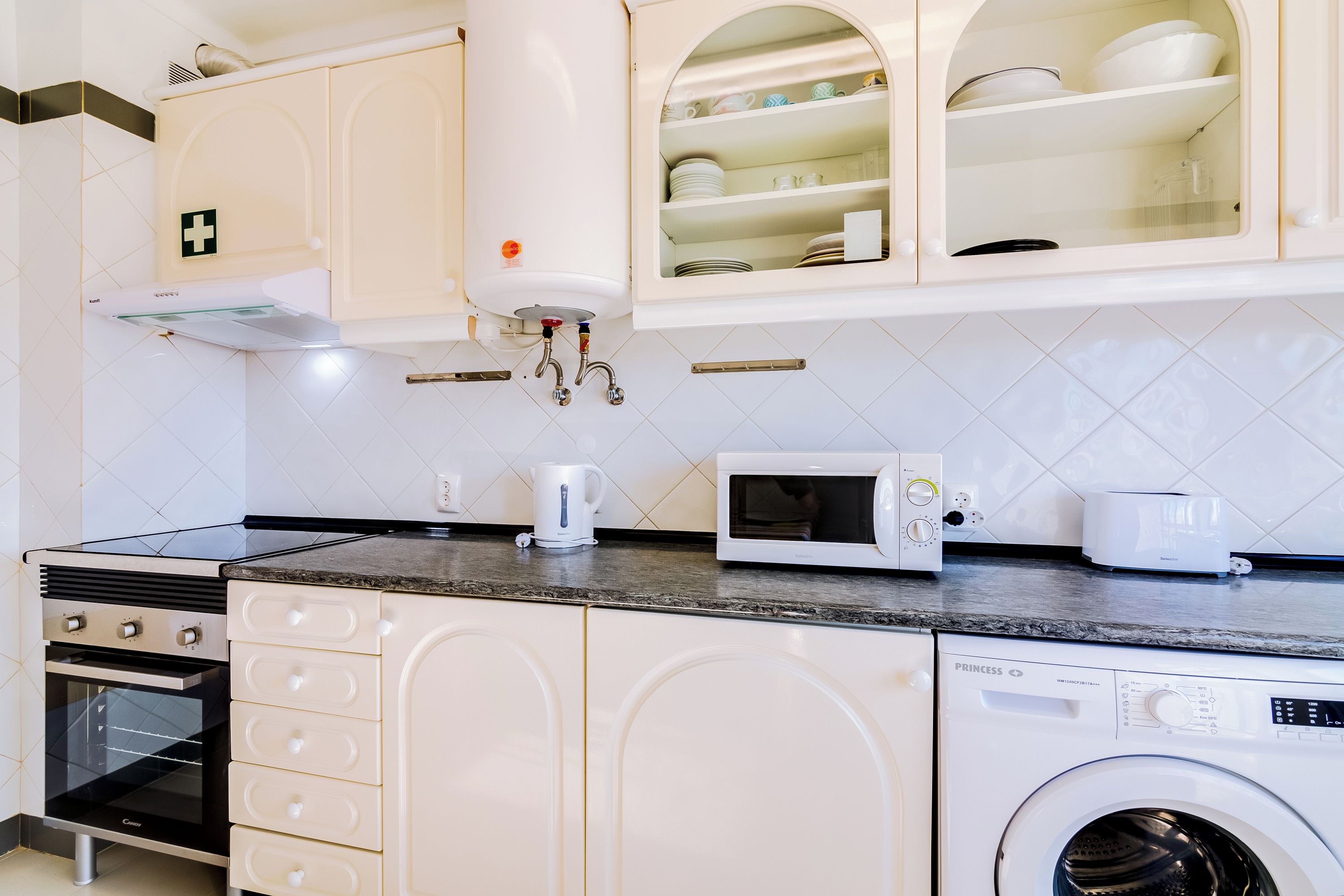 A kitchen with white cabinets, a built-in microwave, a kettle, and a washing machine.