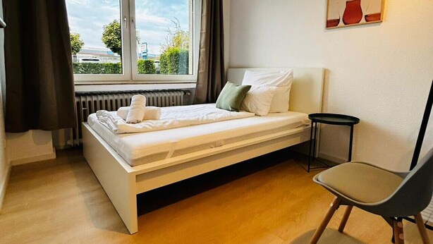 A bedroom with a bed, a chair, a radiator, and a window with curtains. - House Rasink I Fitters I 160m2 (Ibbenbüren)