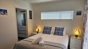 Ein ordentlich eingerichtetes Schlafzimmer mit Bett, Nachttischen, einem Spiegel und einem Fenster mit Jalousien.