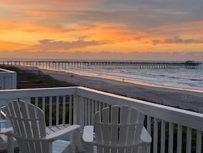 A beach view with a pier extending into the water, a deck with chairs, and a building in the background.