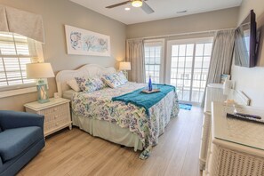 A bedroom with a bed, nightstands, a chair, a TV, and a view through a window. - Ocean Echos – Luxury Oceanfront Vacation Rental in North Topsail Beach (North Topsail Beach)