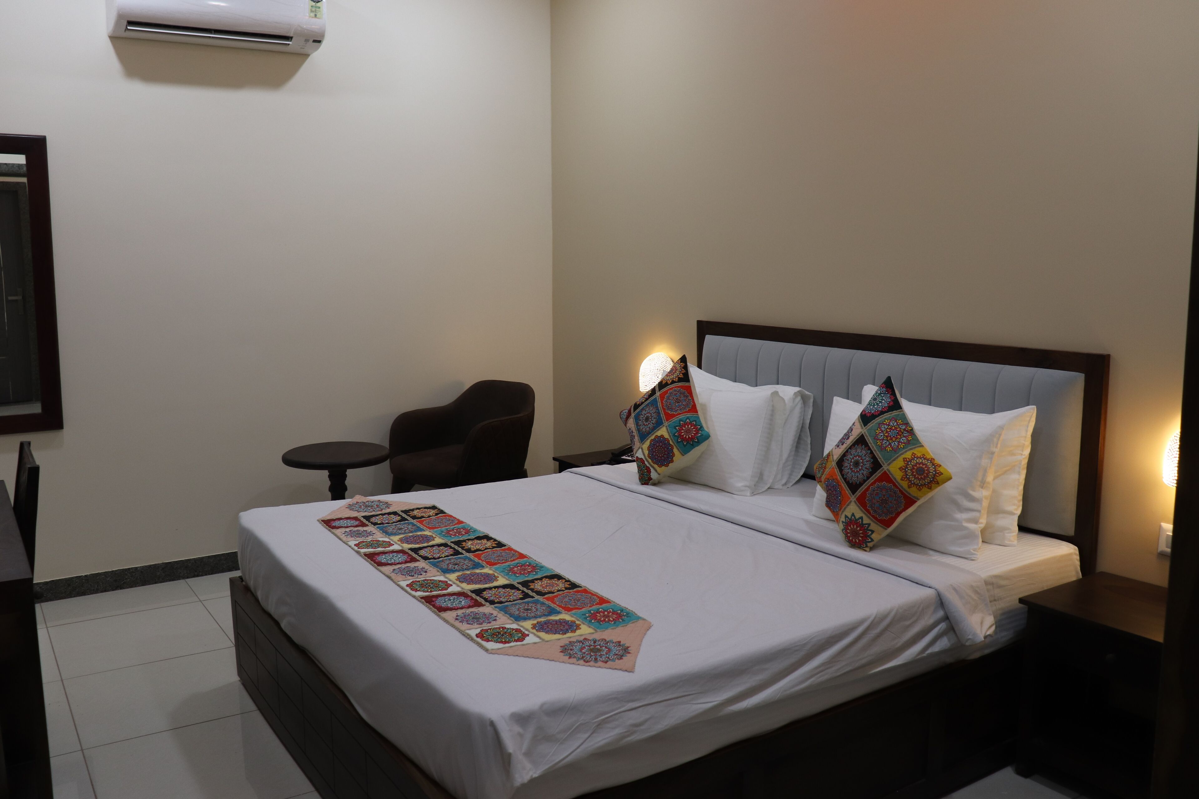 A neatly made bed with a colorful quilt, a bedside table, a chair, and a wall-mounted air conditioner.
