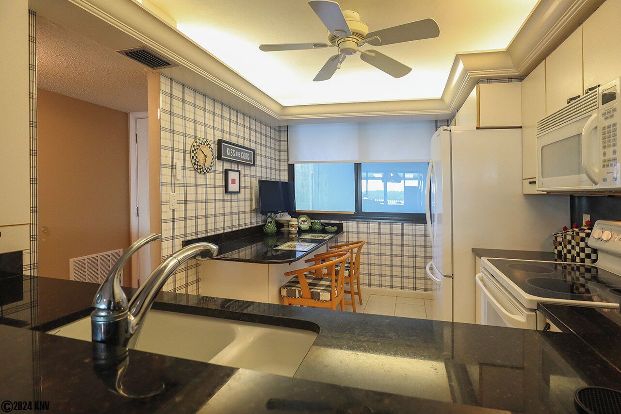 Una cocina con encimera negra, armarios blancos, microondas, estufa y ventilador de techo.