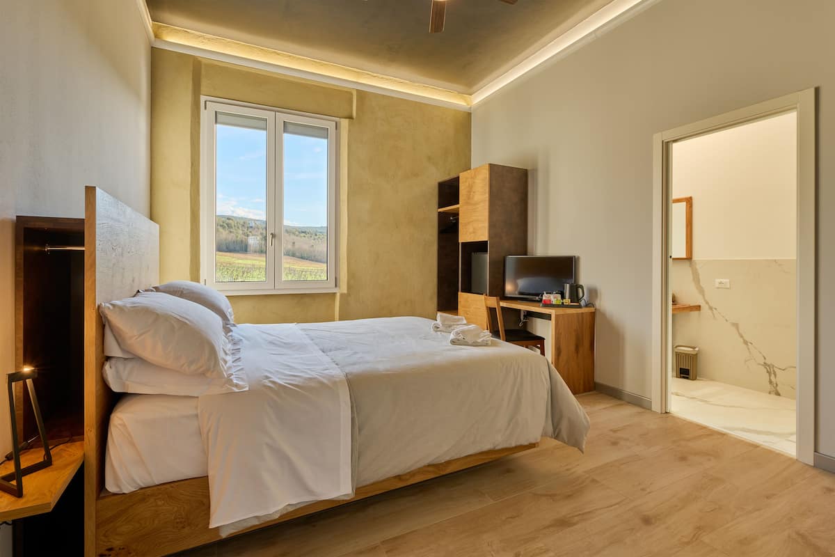 A bedroom with a large bed, a desk with a computer, and a window with a view of the outdoors.