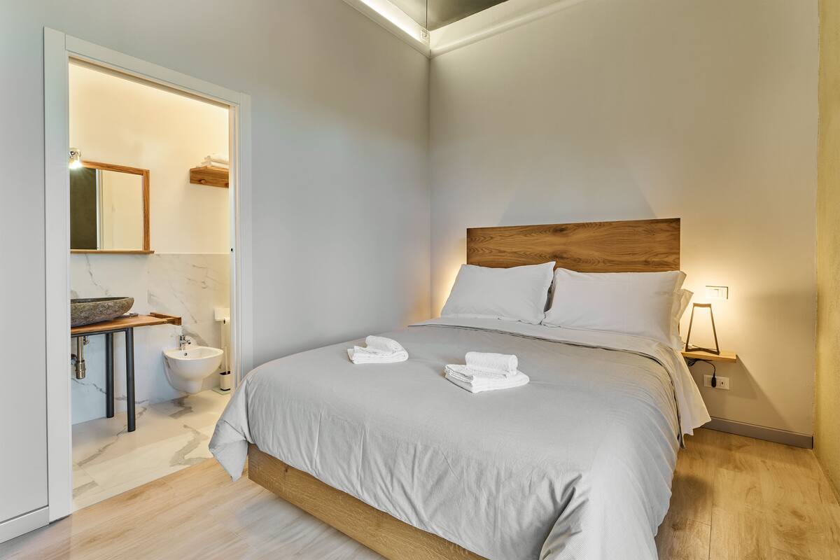 A modern bedroom with a large bed, a wooden headboard, a bedside table with a lamp, and a bathroom with a sink and mirror.