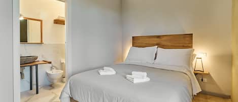 A modern bedroom with a large bed, a wooden headboard, a bedside table with a lamp, and a bathroom with a sink and mirror.