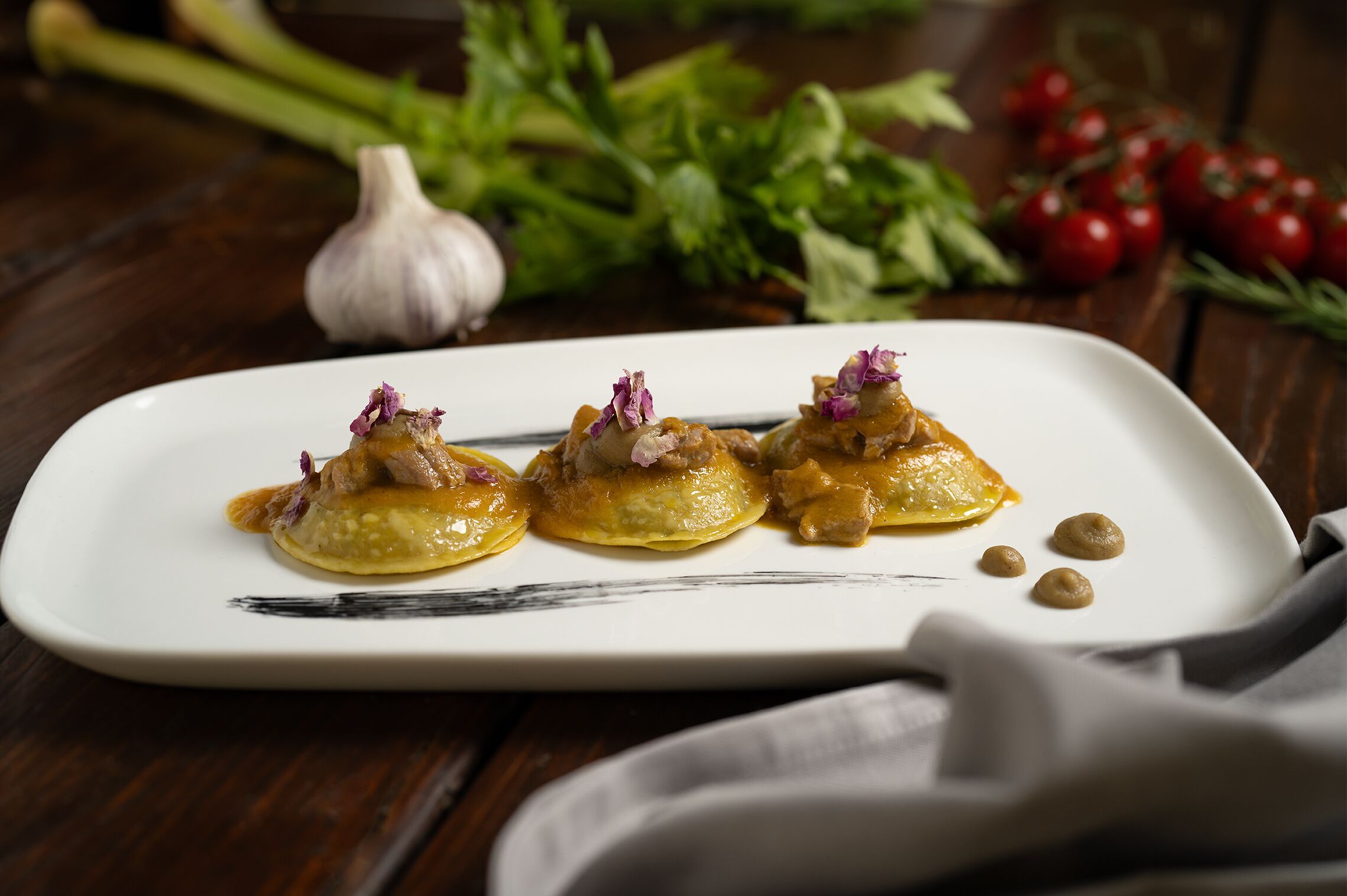 A white rectangular plate with three dumplings topped with a sauce and garnished with purple flowers, placed on a wooden table.
