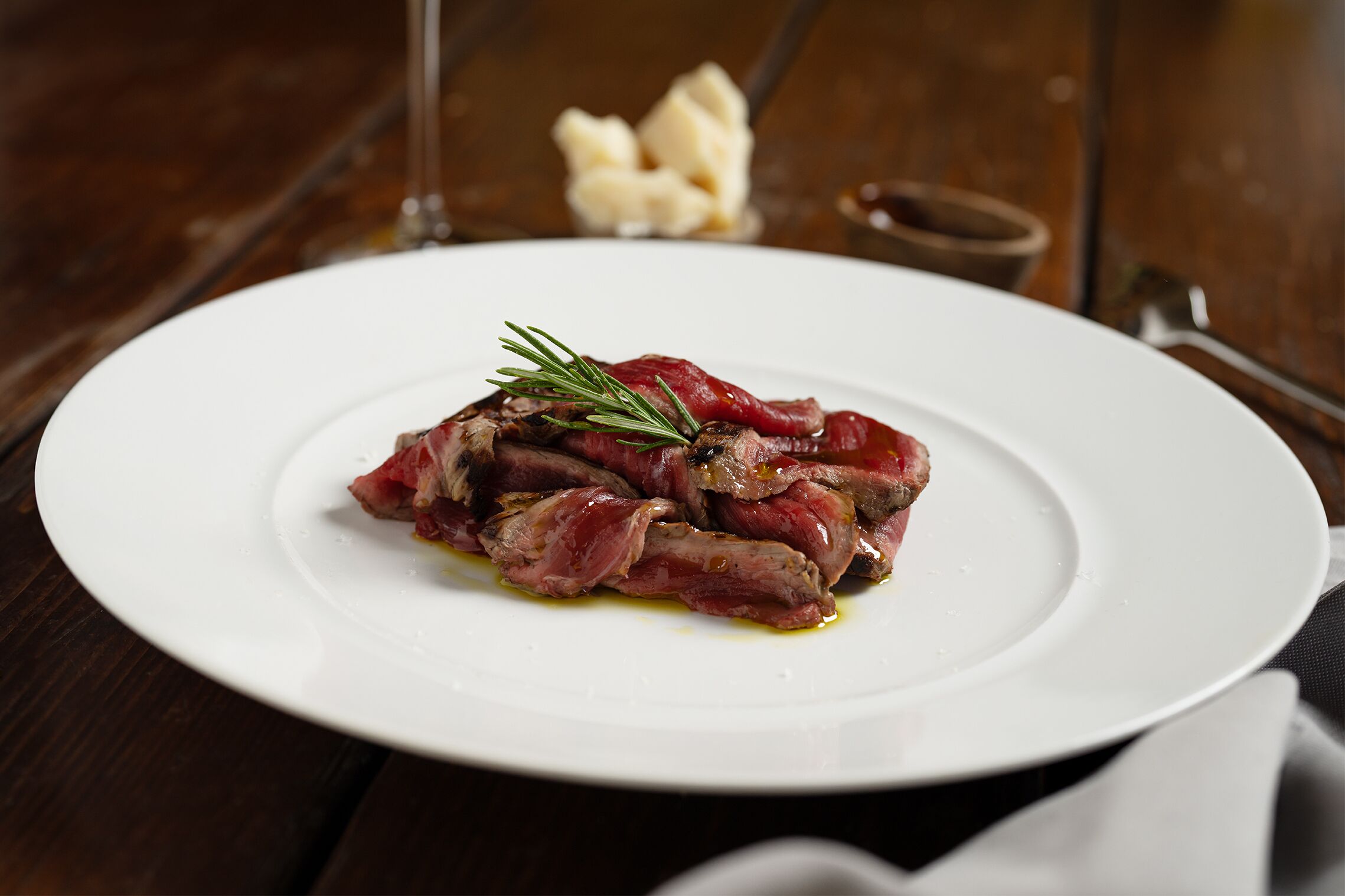 A white plate with sliced meat garnished with rosemary, accompanied by a glass of wine and a small bowl of garlic.