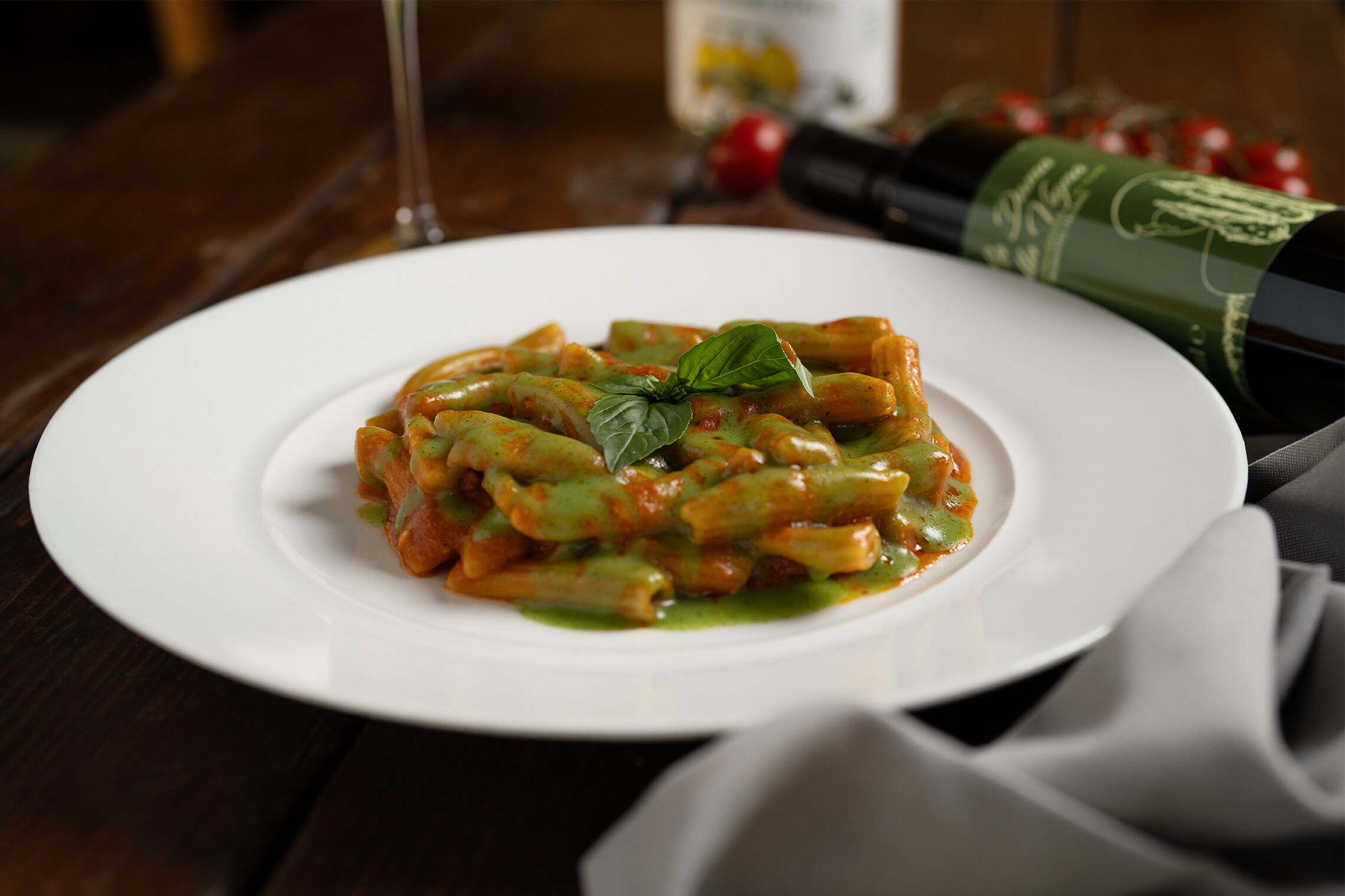 En tallerken med pasta, grøn sauce og et basilikumblad som pynt, samt en vinflaske og et vinglas på et træbord.