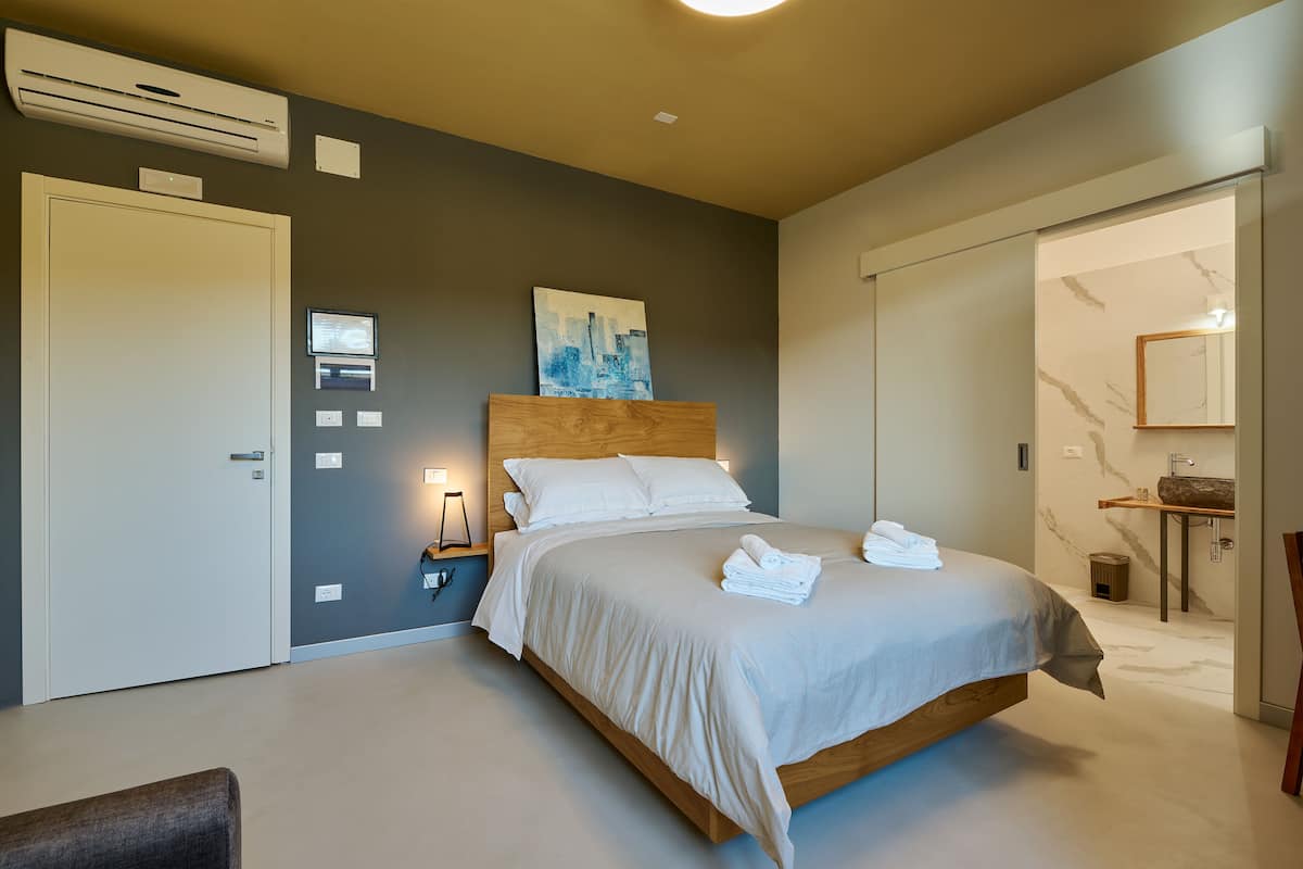 A modern bedroom with a bed, bedside lamps, a cityscape painting, and a bathroom visible through an open door.