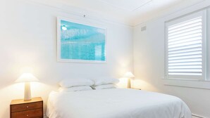 A bedroom with a bed, two bedside tables with lamps, a wall-mounted painting, and a window with blinds.