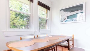 A dining area with a long wooden table, white chairs, and a bench. There are two windows with blinds, and a framed picture on the wall.