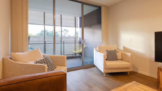 Una sala moderna con sofá, sillón y una pequeña mesa de madera. Hay una puerta corrediza de vidrio que da a un balcón con una silla afuera.