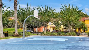 An outdoor basketball court with a hoop, surrounded by palm trees and a yellow building in the background. - Lakeview Home in Terra Verde:Pool & Resort Amenities (Kissimmee)