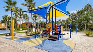 A playground with a blue and yellow play structure, surrounded by palm trees and a paved area. - Lakeview Home in Terra Verde:Pool & Resort Amenities (Kissimmee)
