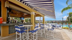 An outdoor bar area with striped awnings, seating, and a pool in the background. - Lakeview Home in Terra Verde:Pool & Resort Amenities (Kissimmee)