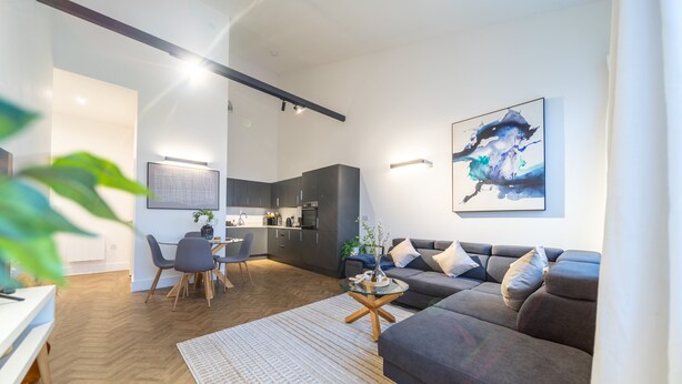 A modern living room with a grey sofa, a dining area with a table and chairs, and a kitchen in the background.