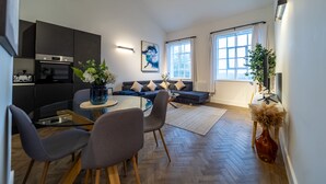 A modern living room with a sofa, a dining table, and chairs. There is a kitchen area with cabinets and appliances.