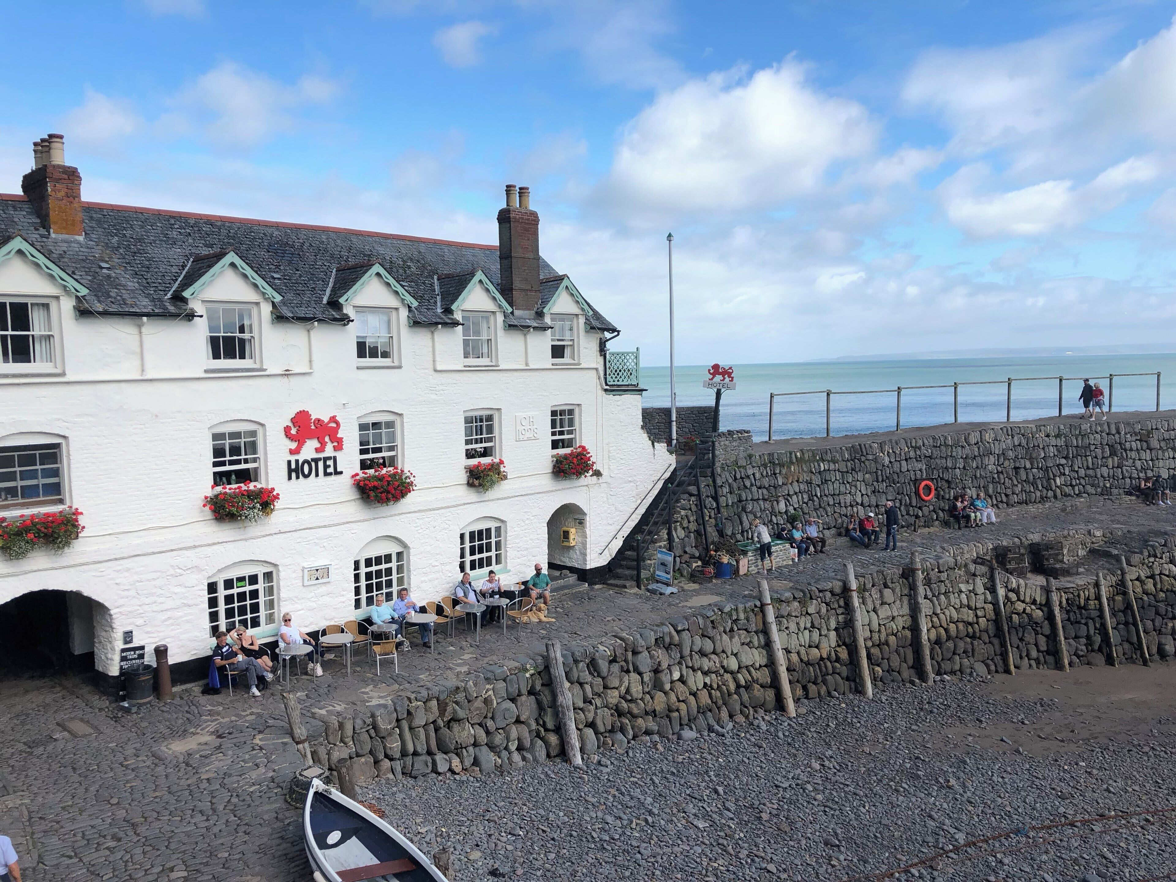 A white building with a sign reading "HOTEL" and a lion emblem. There are flower boxes on the windows, and a stone wall in front.