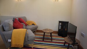 Ein Wohnzimmer mit grauem Sofa, roten und gelben Kissen, einem kleinen Holztisch, einer TV-Kommode und einem gestreiften Teppich.