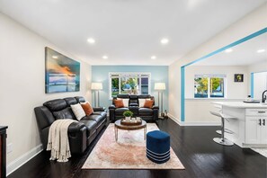 A modern living room with a black leather sofa, a coffee table with a plant, and a blue ottoman. There are two windows with views of a garden and a white kitchen area with a sink and cabinets. - Fenced Dog Yard, 3 Full Baths, Sleeps 8, Minutes to Downtown (Lake Geneva)