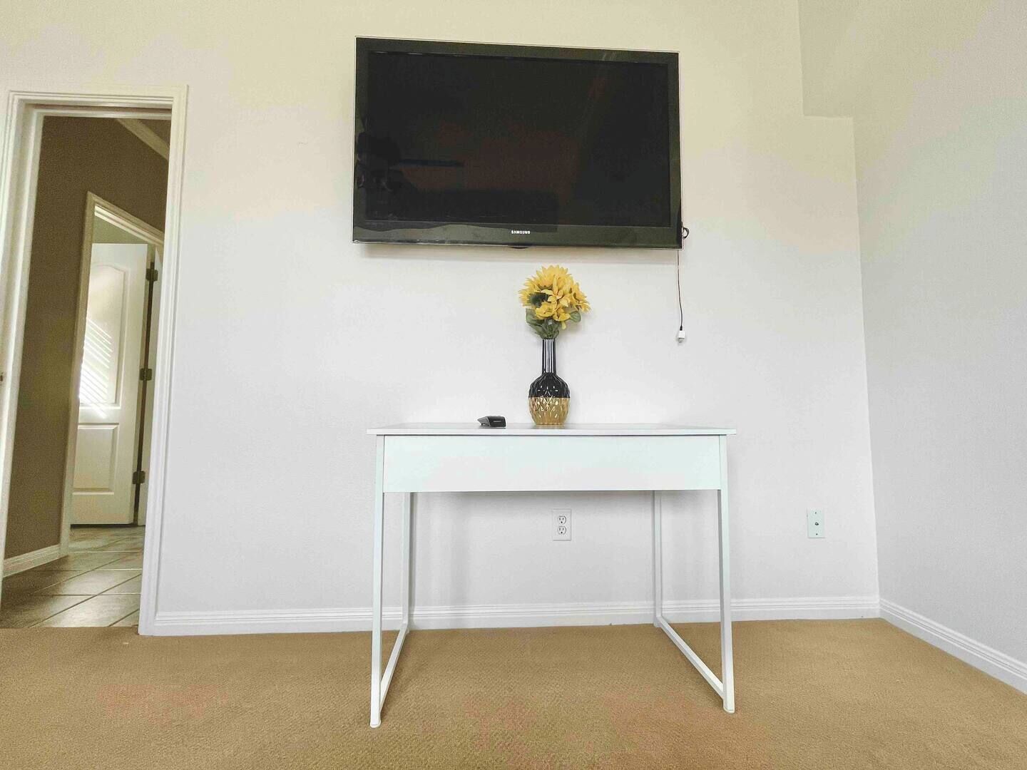 A room with a white wall, a mounted TV, a white console table with a vase of flowers, and a door leading to another room.