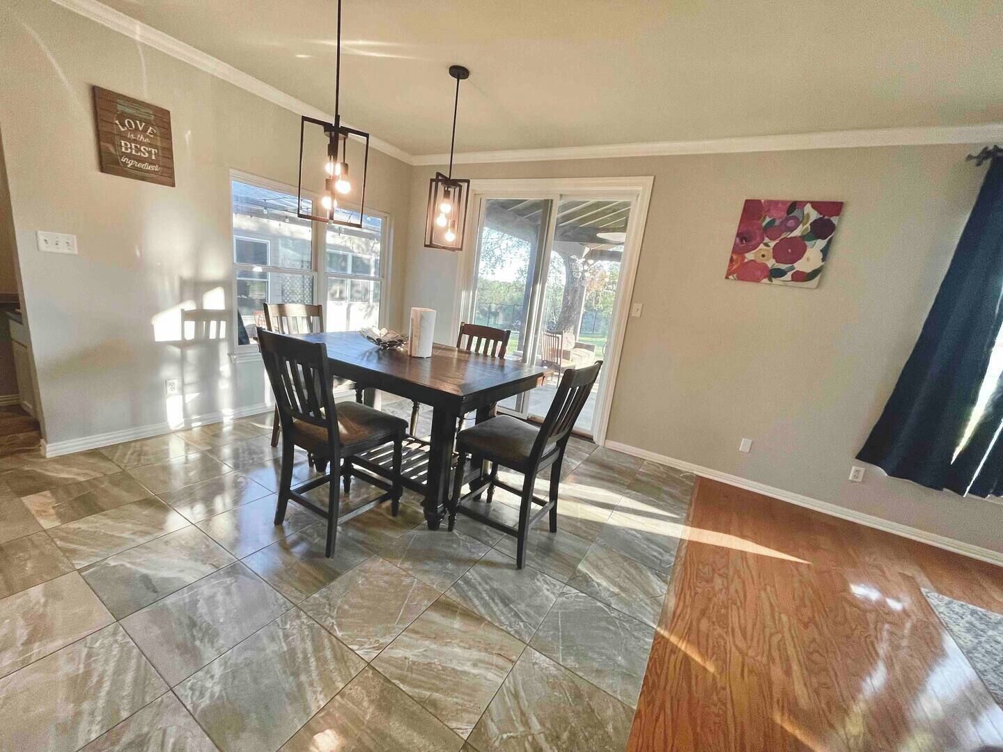 A dining area with a dark wooden table and chairs, a hanging light fixture, and a kitchen visible through an open door.