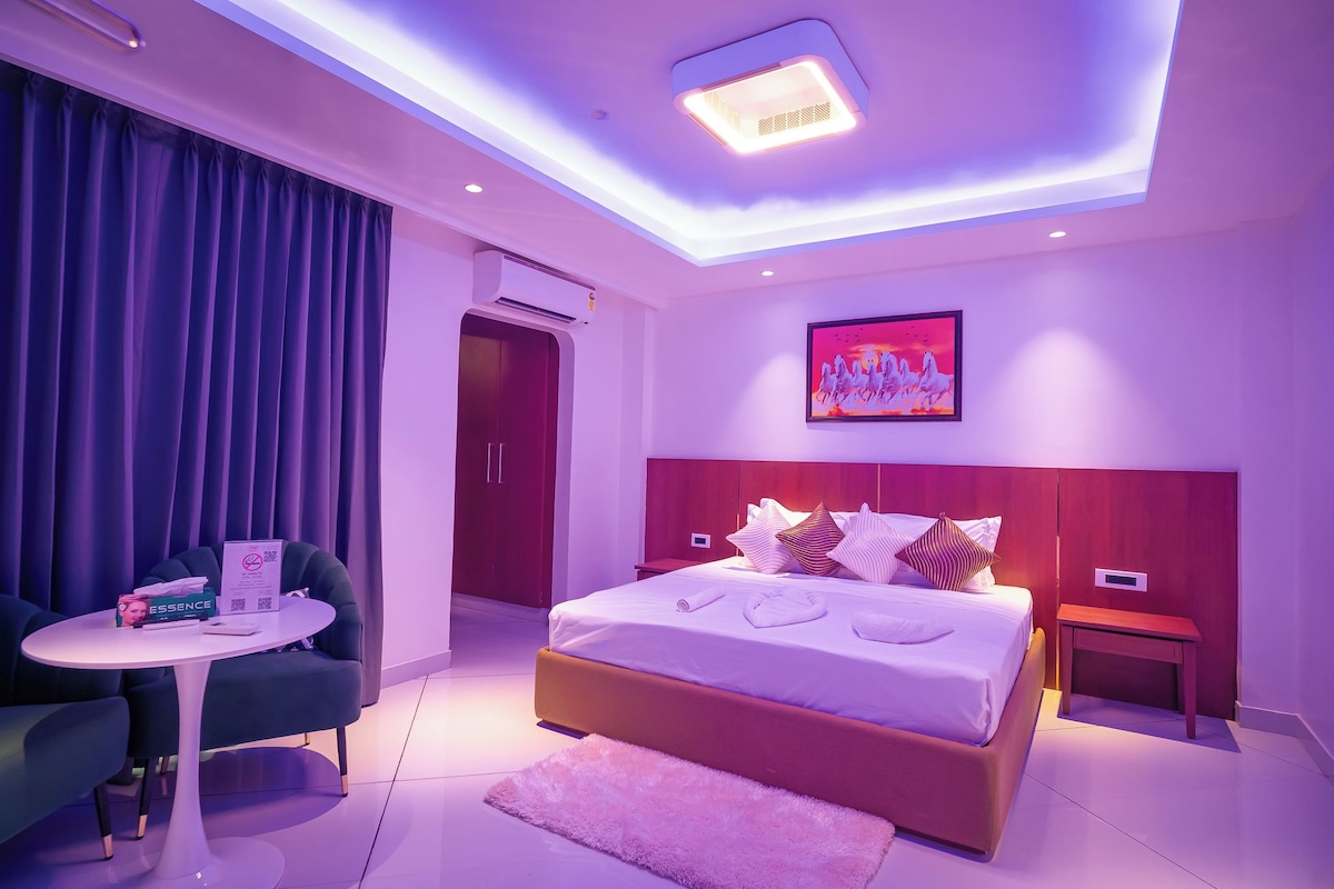 A modern bedroom with a bed, bedside tables, a small table with a chair, a ceiling light, and a painting on the wall.