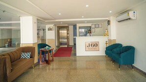 A hotel reception area with a reception desk labeled "J60 SUITES," a brown sofa, teal armchairs, a small table with a red rug, and an elevator in the background. - J60 Suites (New Delhi)