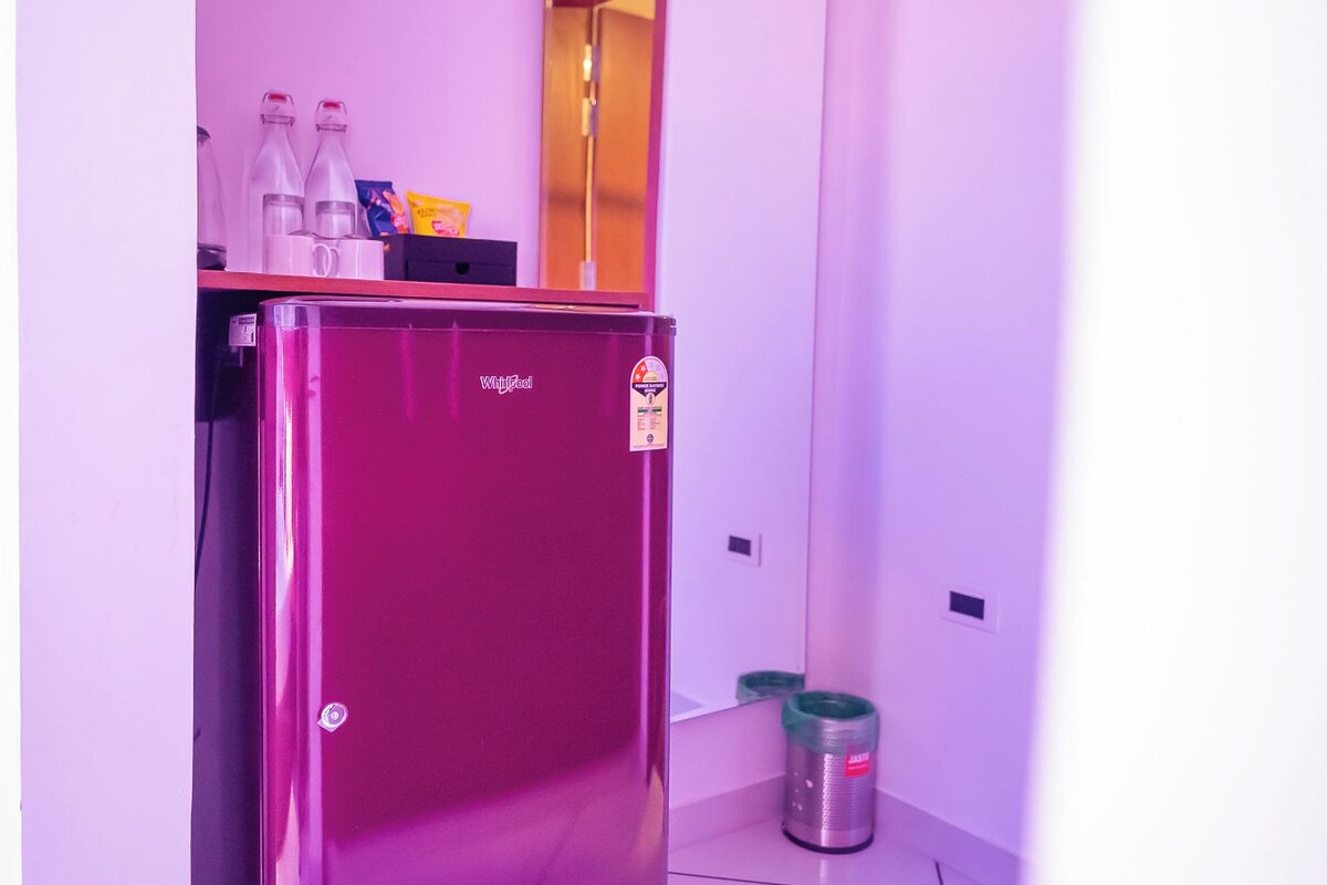 A small, modern kitchen with a pink refrigerator, a trash can, and a countertop with bottles and a box.