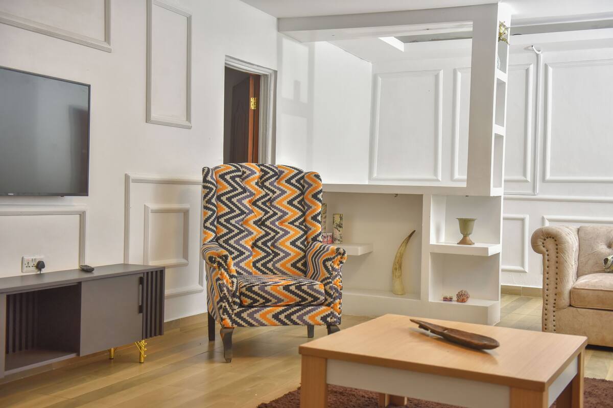 Een woonkamer met een modern design, met een grote flatscreen-tv, een gestreept fauteuil, een houten salontafel en een beige bank.