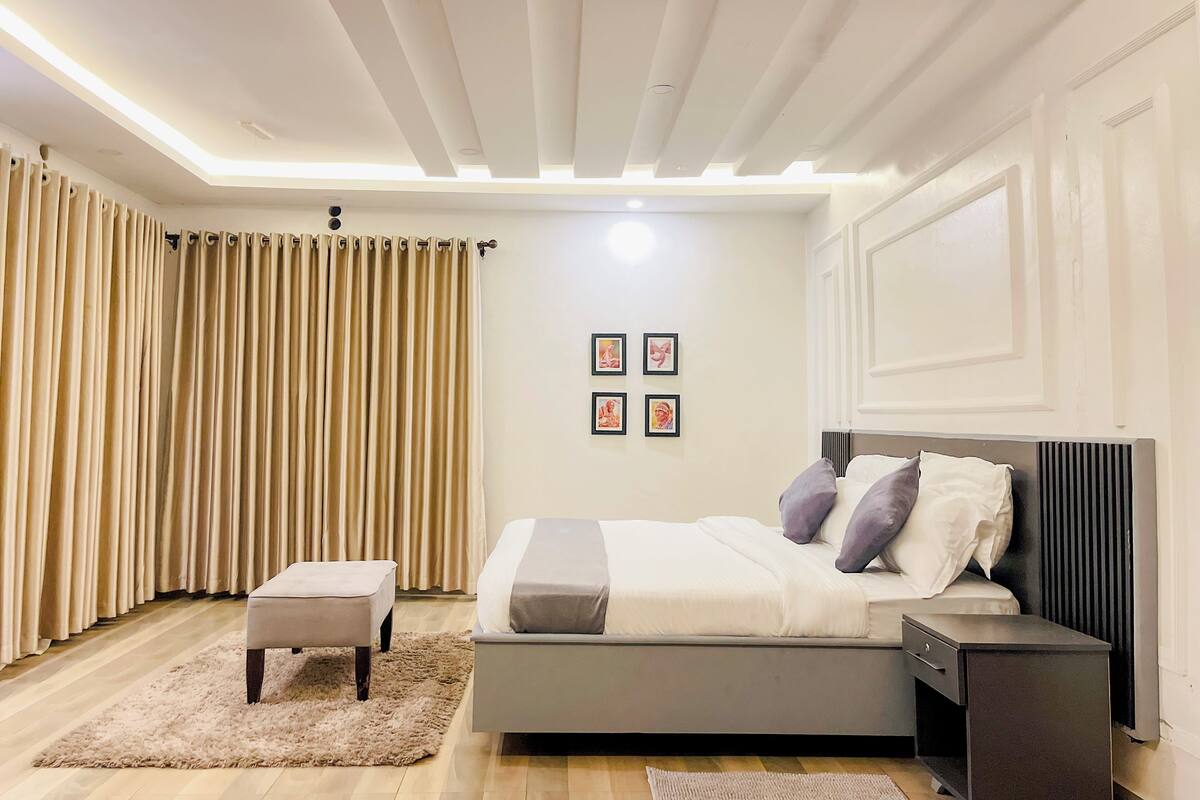 Een moderne slaapkamer met een bed, nachtkastjes, een klein krukje en een plafond met ingebouwde verlichting.