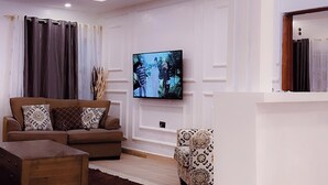 A modern living room with a sofa, a coffee table, and a patterned armchair. A flat-screen TV is mounted on the wall. - Olakira Mara Homes (Maasai Mara)