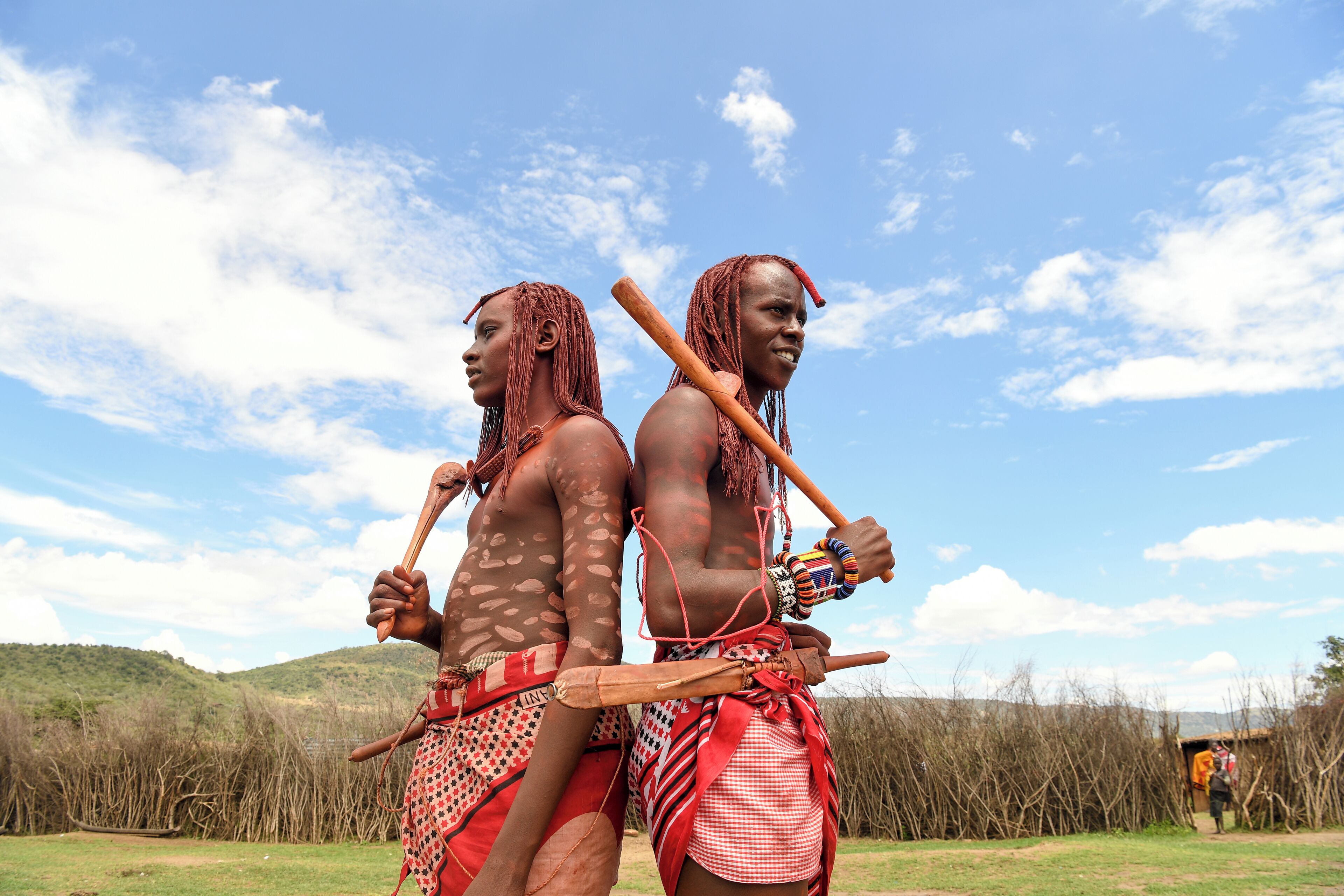 Two individuals in traditional attire with wooden staffs, standing outdoors with a clear sky and distant hills.