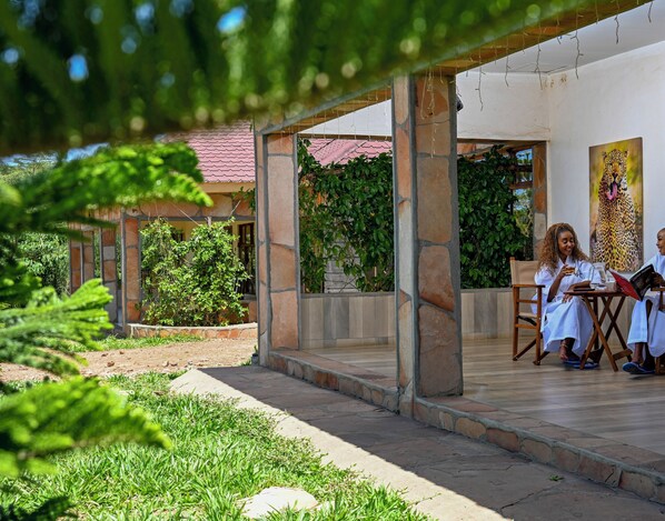 Un espacio al aire libre cubierto con pilares de piedra, piso de baldosas y vista a una casa con techo rojo y vegetación.