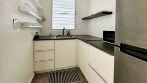 Une cuisine de taille réduite avec des meubles blancs, un plan de travail noir, un réfrigérateur en acier inoxydable et une fenêtre équipée de stores.