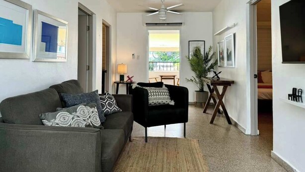 Un vaste espace de vie comprenant un canapé gris, un fauteuil noir et une table à manger avec des chaises. On y trouve également un ventilateur de plafond et une grande fenêtre qui laisse entrer la lumière naturelle.