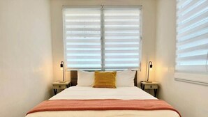Un lit bien fait, recouvert d’une couvre-lit blanche et orange, avec deux lampes de chevet et une fenêtre équipée de stores.
