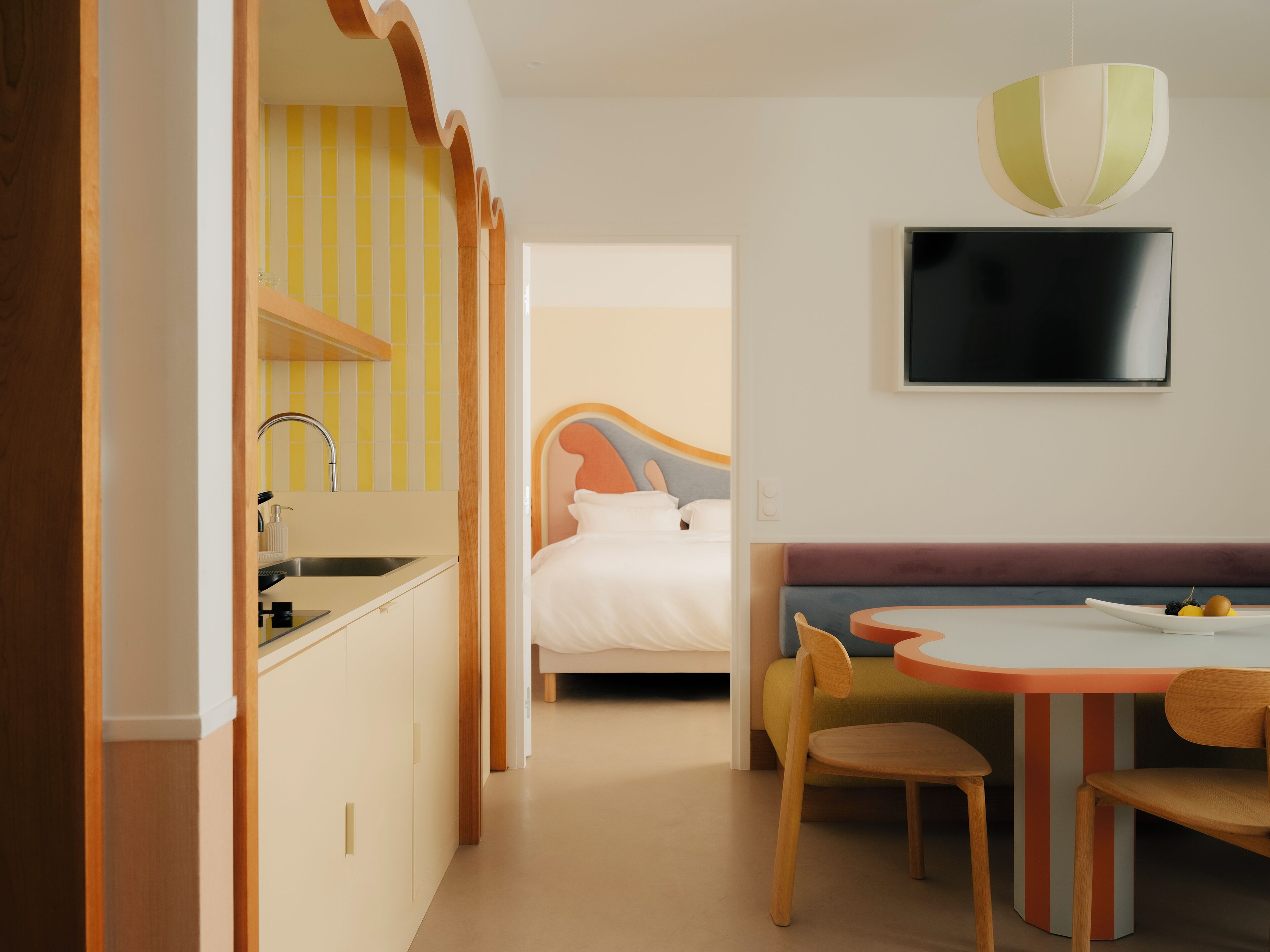 A modern hotel room with a kitchenette, a bed with a colorful headboard, a dining area with a table and chairs, and a flat-screen TV mounted on the wall.