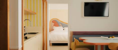 A modern hotel room with a kitchenette, a bed with a colorful headboard, a dining area with a table and chairs, and a flat-screen TV mounted on the wall.