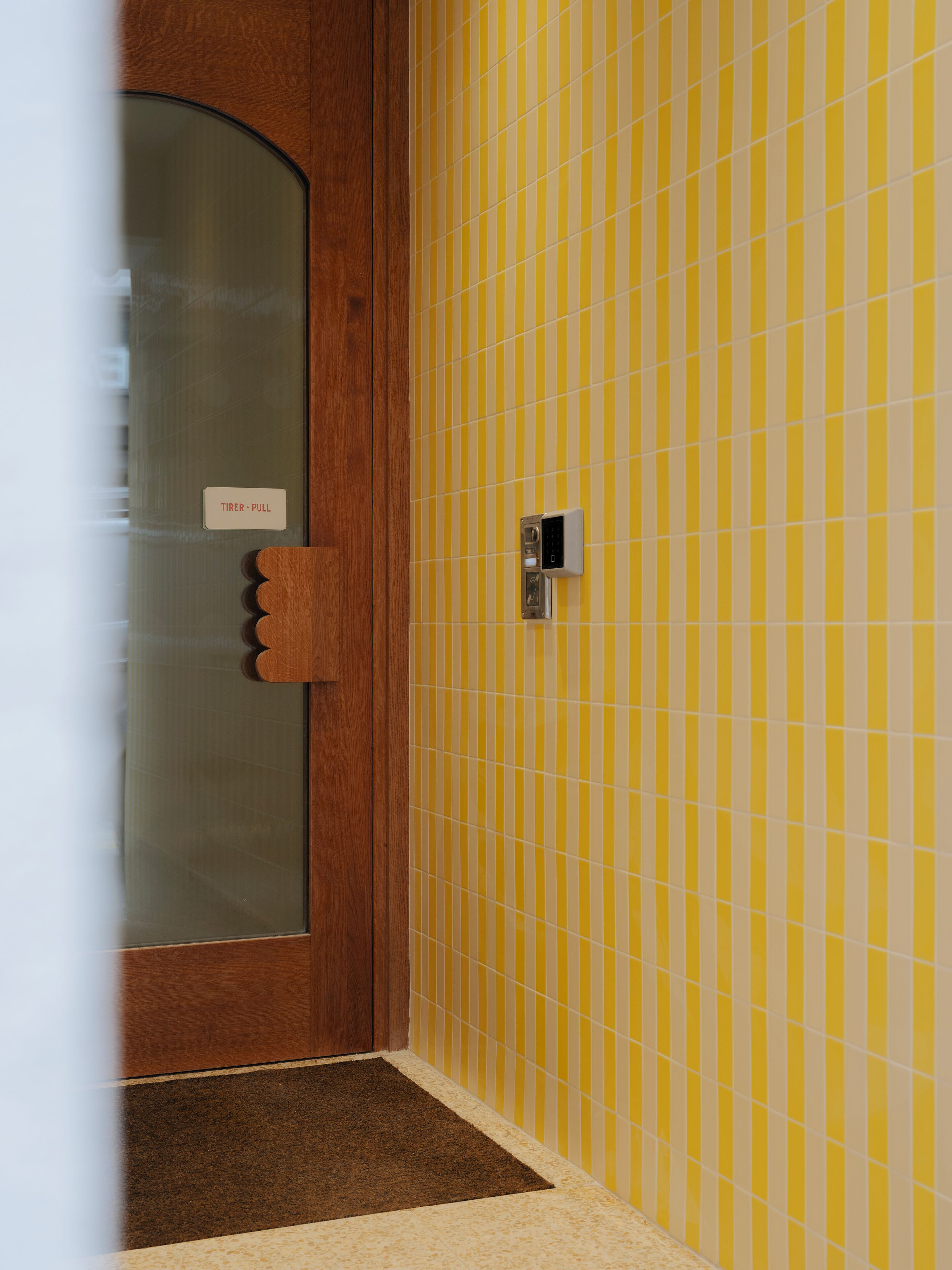 Eine Tür mit Glasscheibe und einem Schild mit der Aufschrift 'TIMER - PULL', eine Wand mit gelb-weiß gestreiften Fliesen und eine braune Fußmatte.