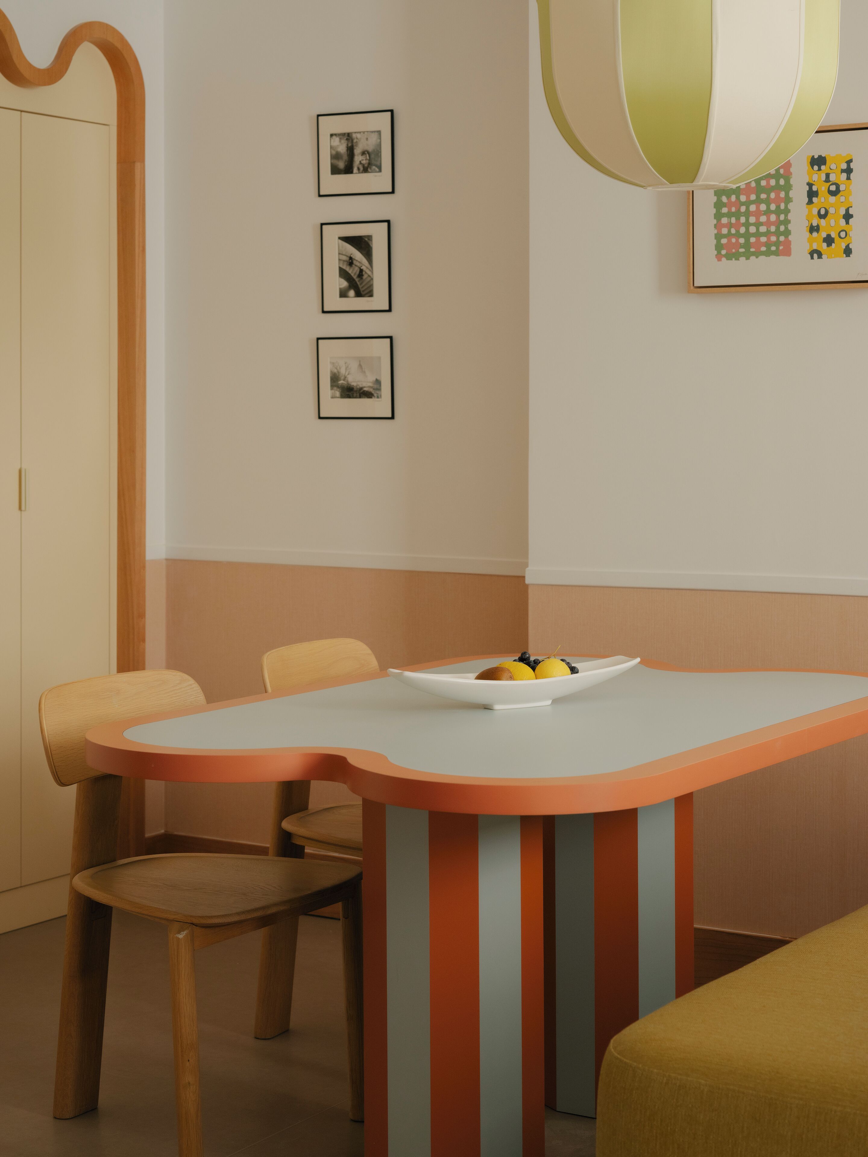A dining area with a table and chairs, a bowl of fruit, and framed pictures on the wall.