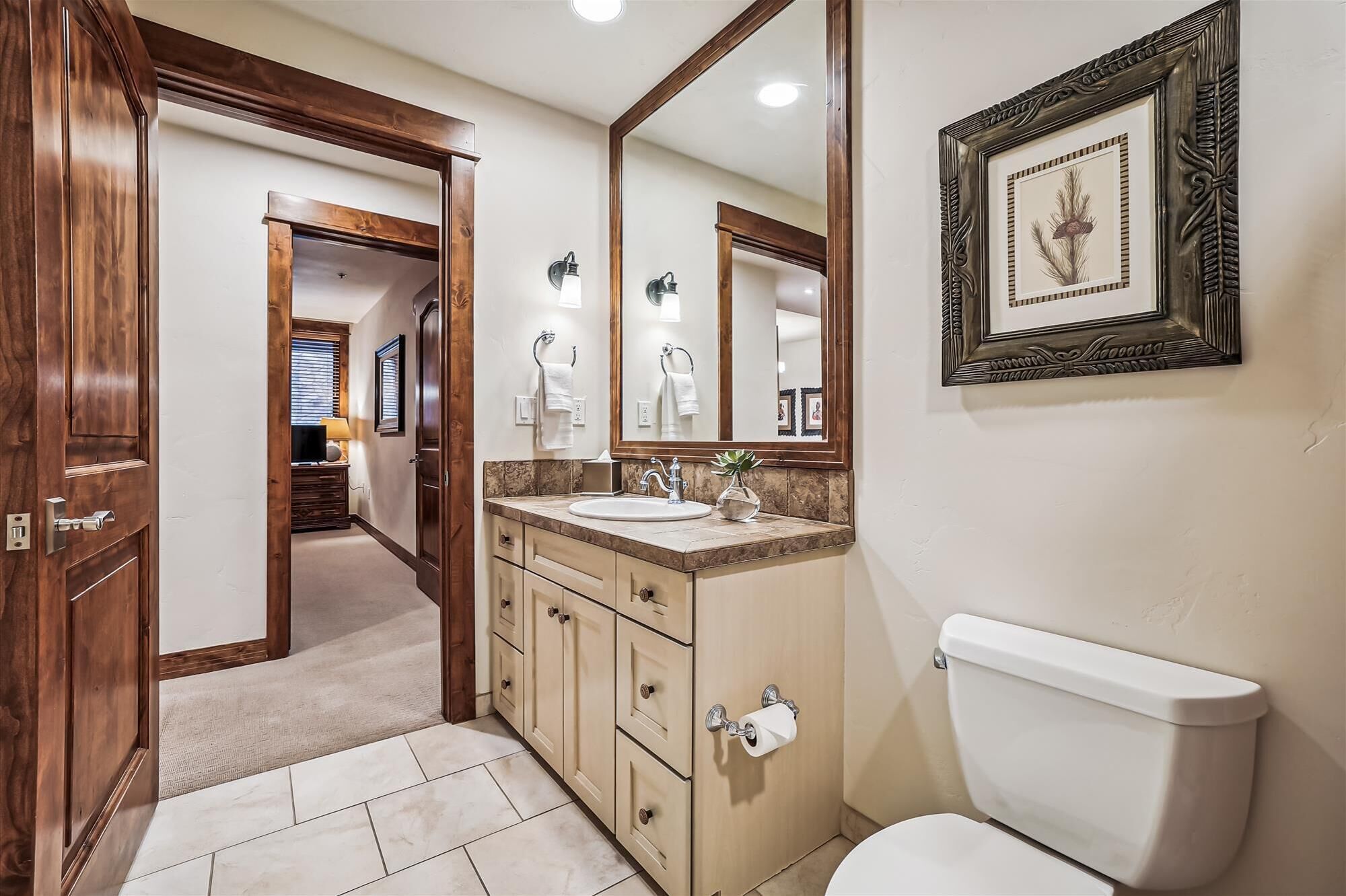 A bathroom with a toilet, sink, and mirror. There is a framed picture on the wall.